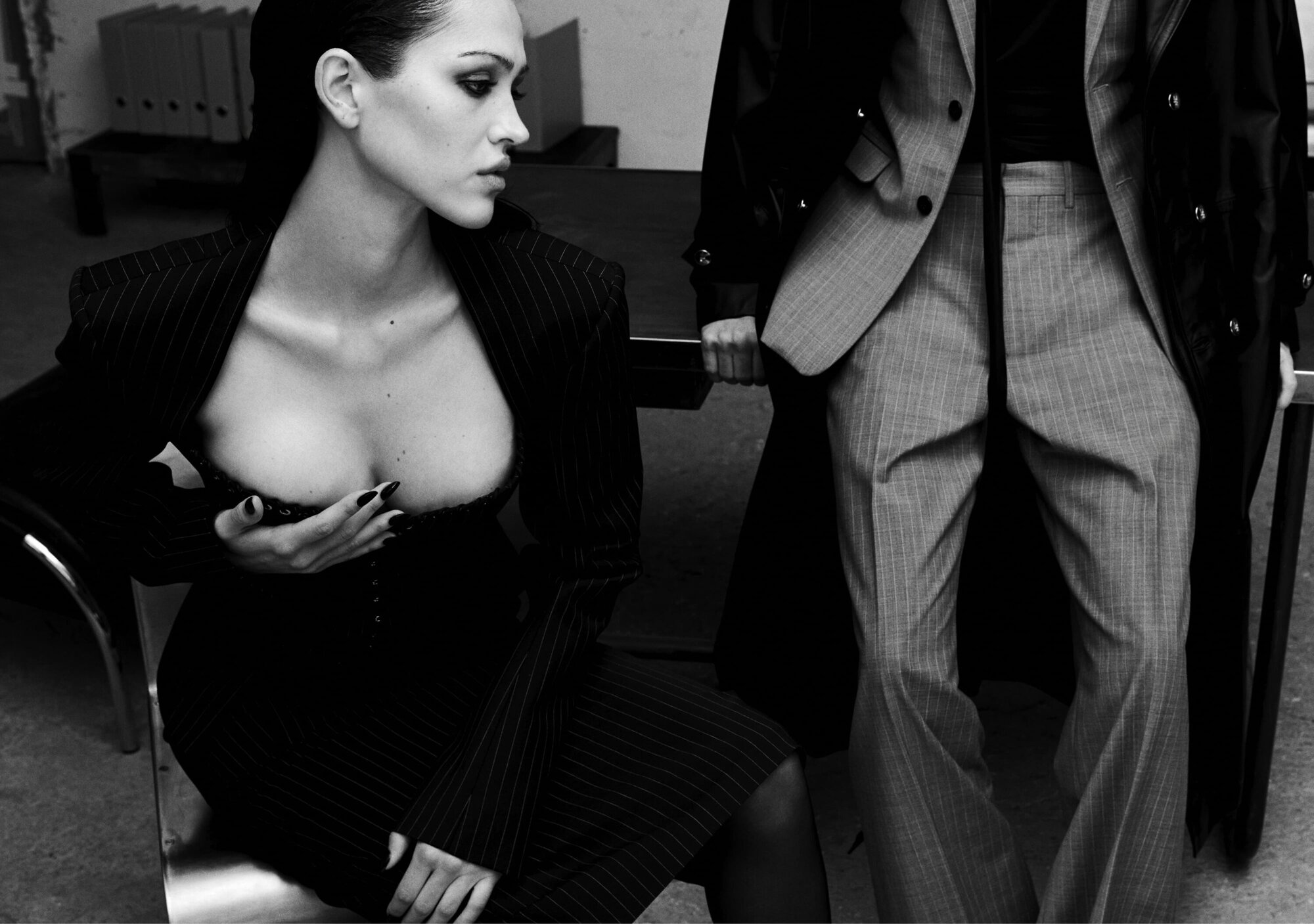 Black and white editorial fashion portrait of a seated woman in a pinstripe jacket next to a standing person in trousers.