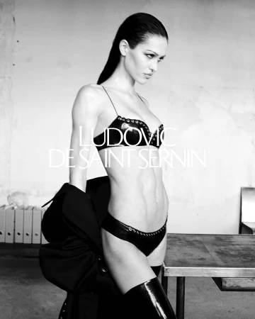 Model in dark undergarments and thigh-high boots with slicked hair standing in an interior office setting.