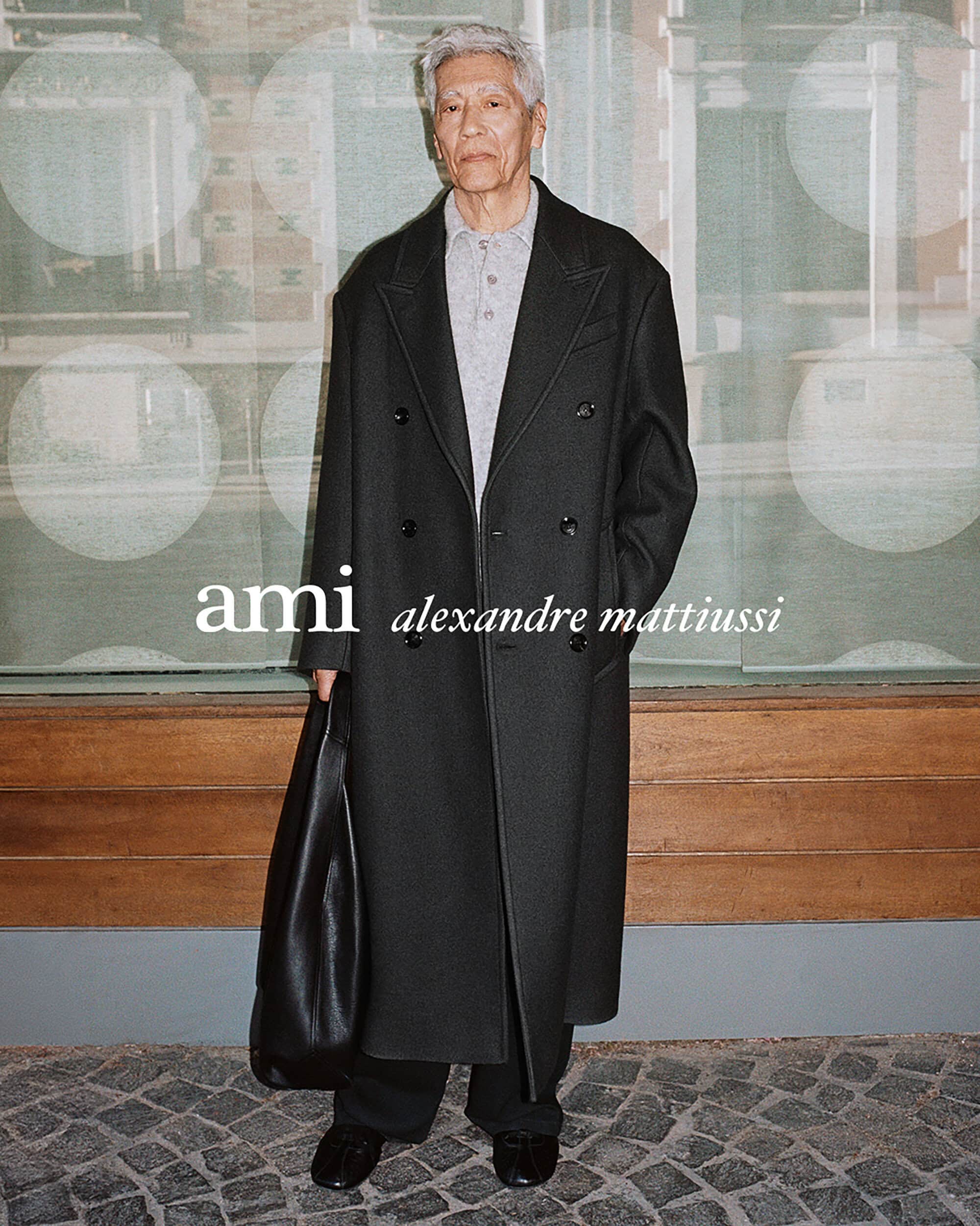 Elderly man wearing a long dark coat holding a large black leather bag standing before a patterned storefront