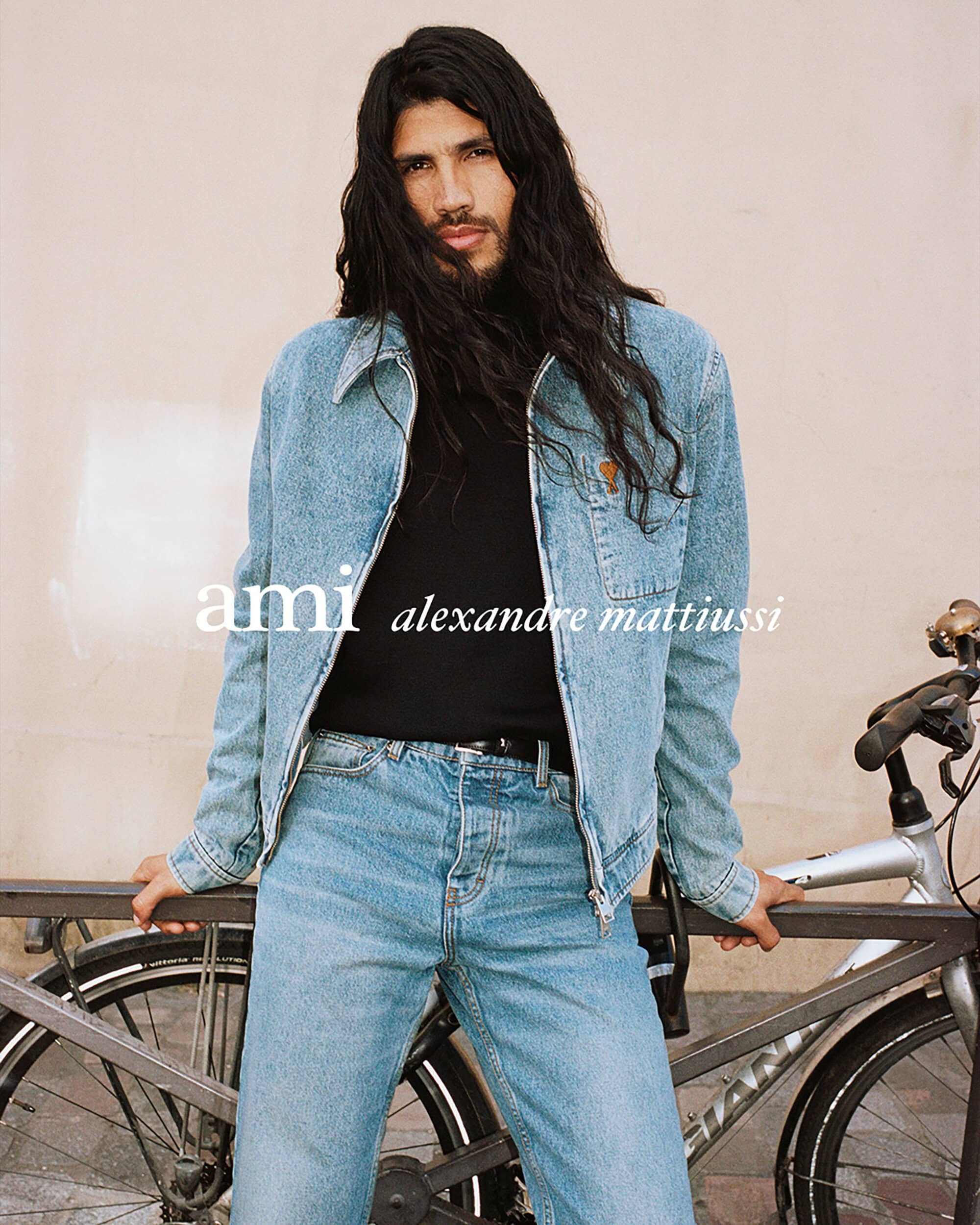 Man with long dark hair wearing a light blue denim jacket and matching jeans leaning against a bicycle