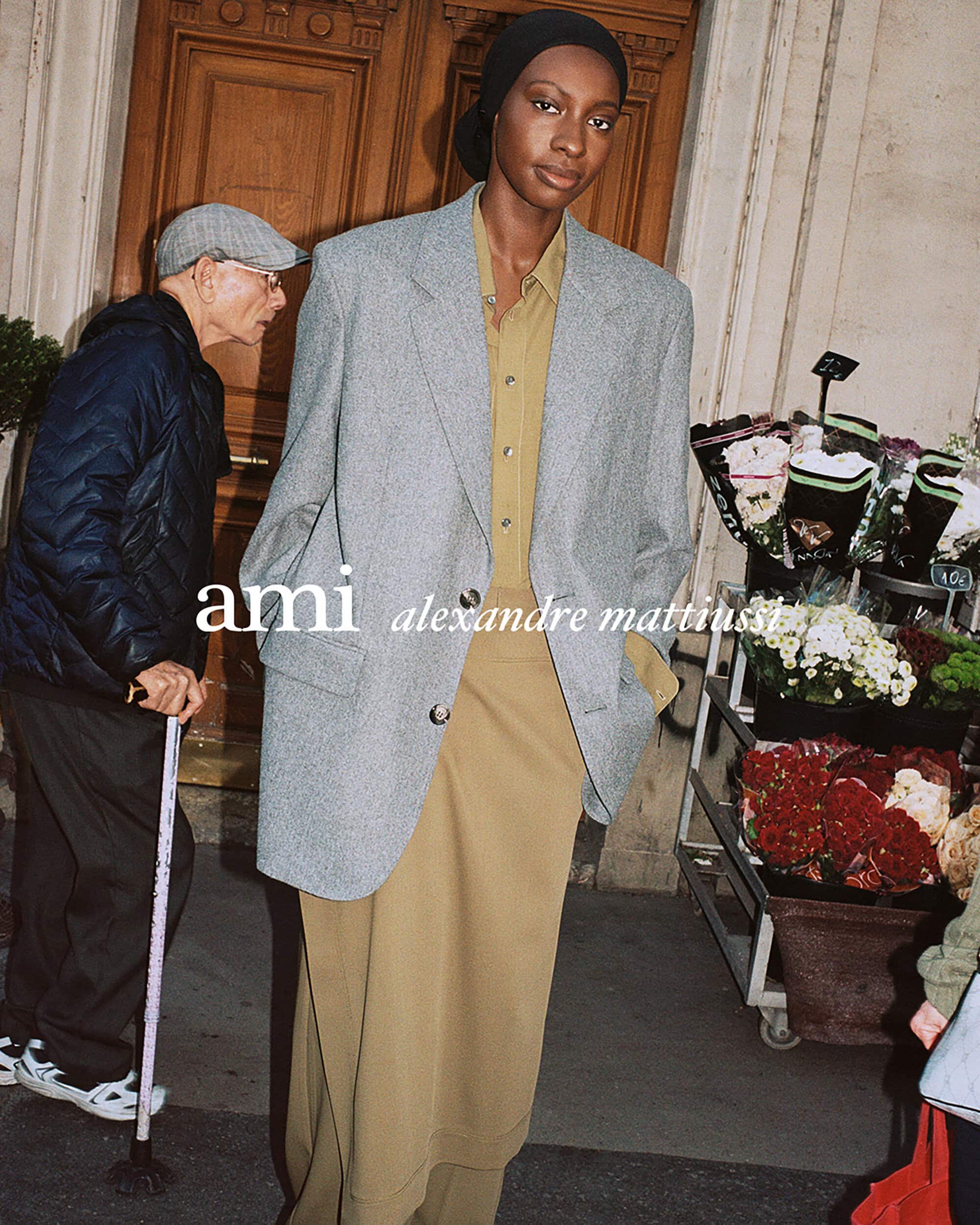 Model in grey blazer and khaki tunic standing on a city street beside a flower cart for Ami Fall 2025 ad campaign.