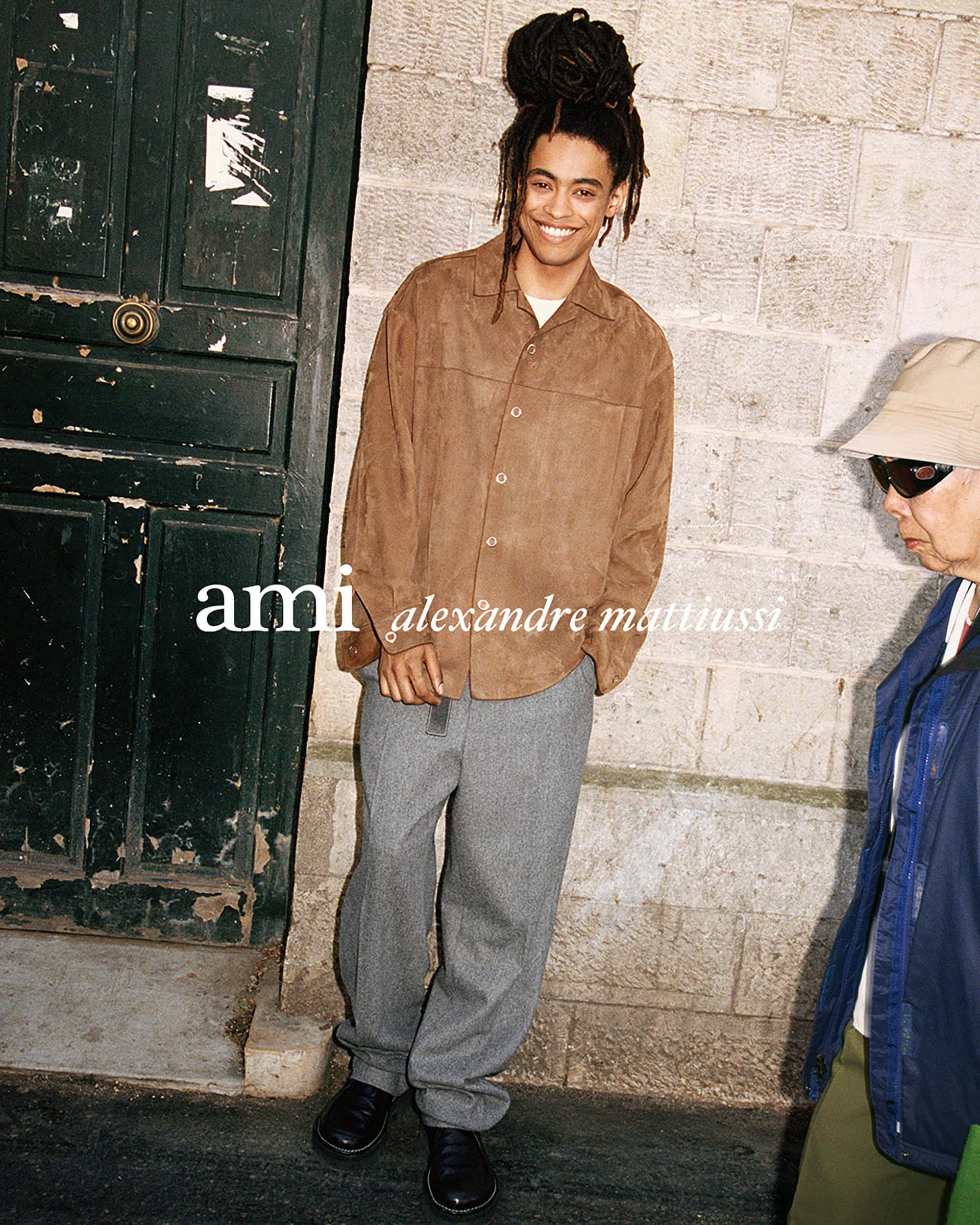 Young smiling man with dreadlocks wearing a brown button-up shirt and grey trousers standing outdoors against a stone wall