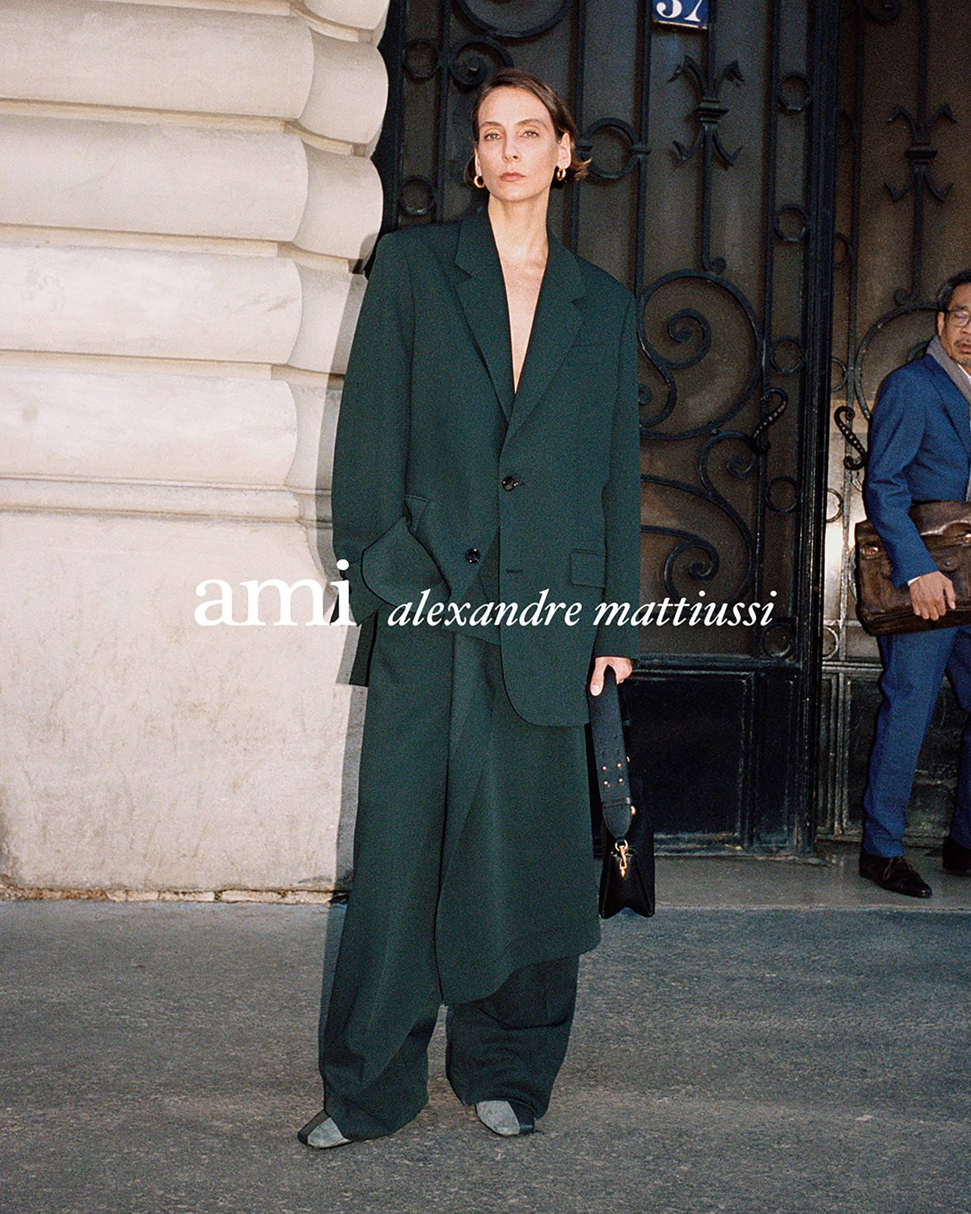Model wearing a dark green oversized blazer and matching trousers holding a handbag in front of a stone building exterior.