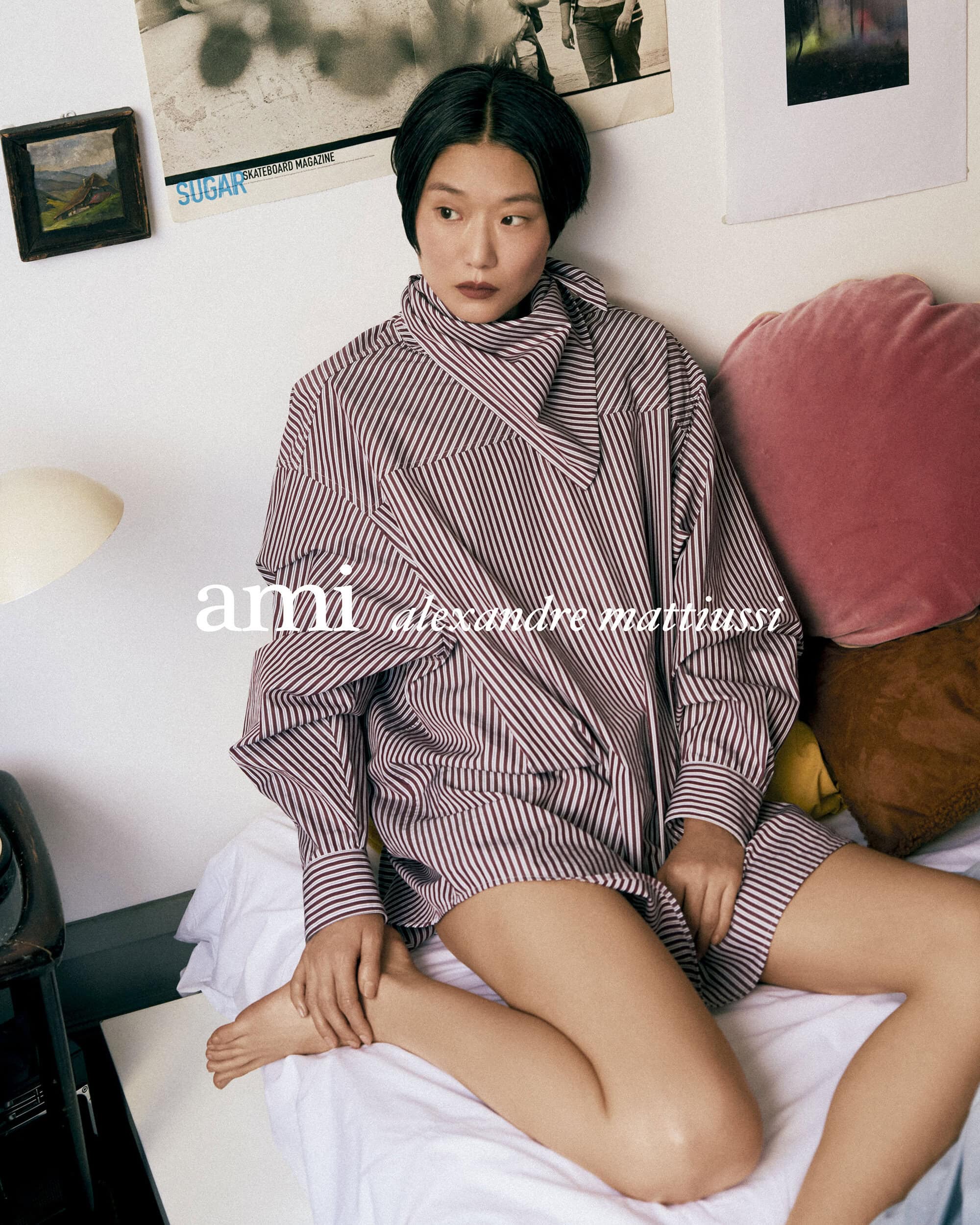 Model seated on a bed wearing a striped oversized shirt with a matching neck wrap for Ami