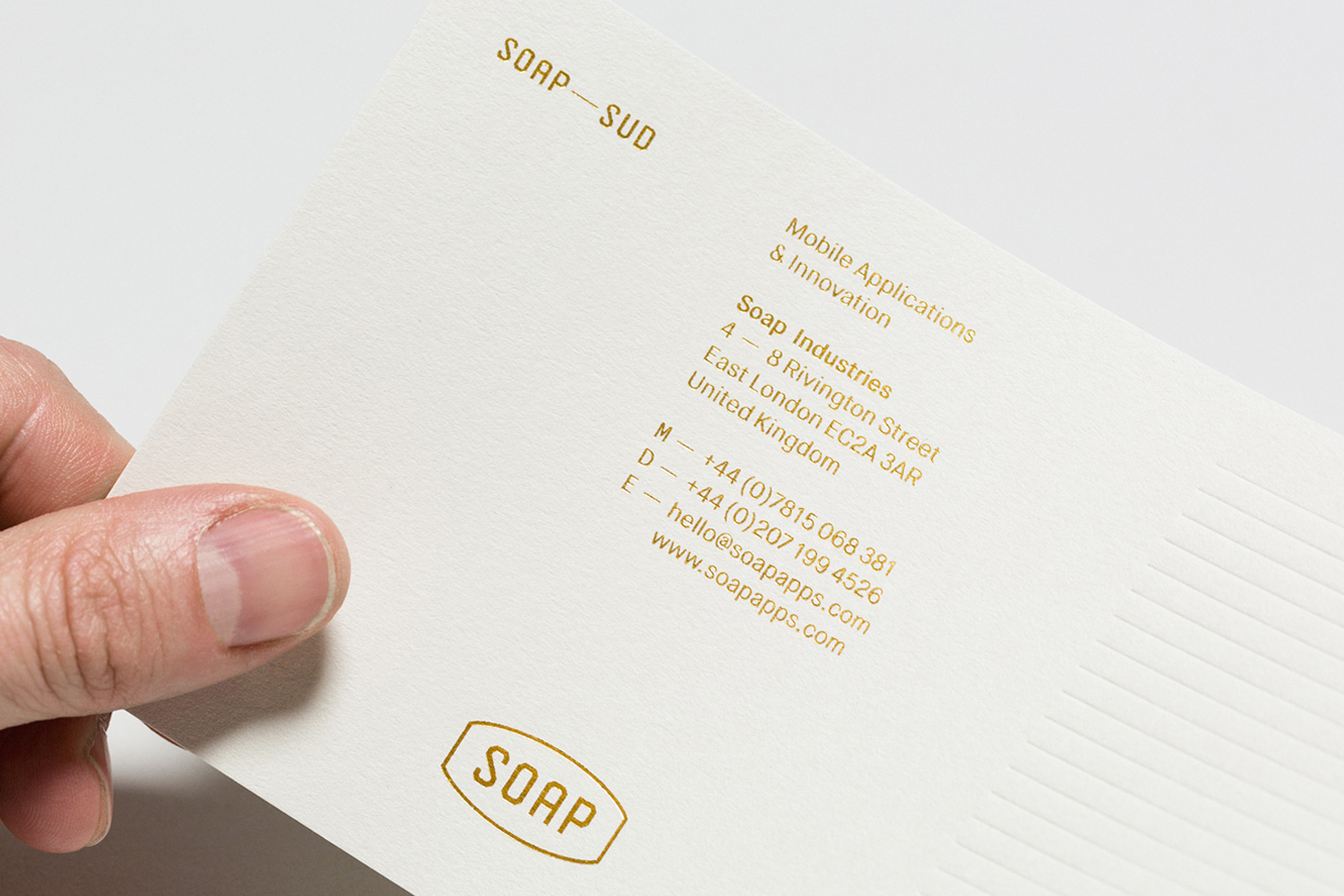 A hand holding a cream textured paper postcard featuring gold foil embossed typography for Soap Industries branding.