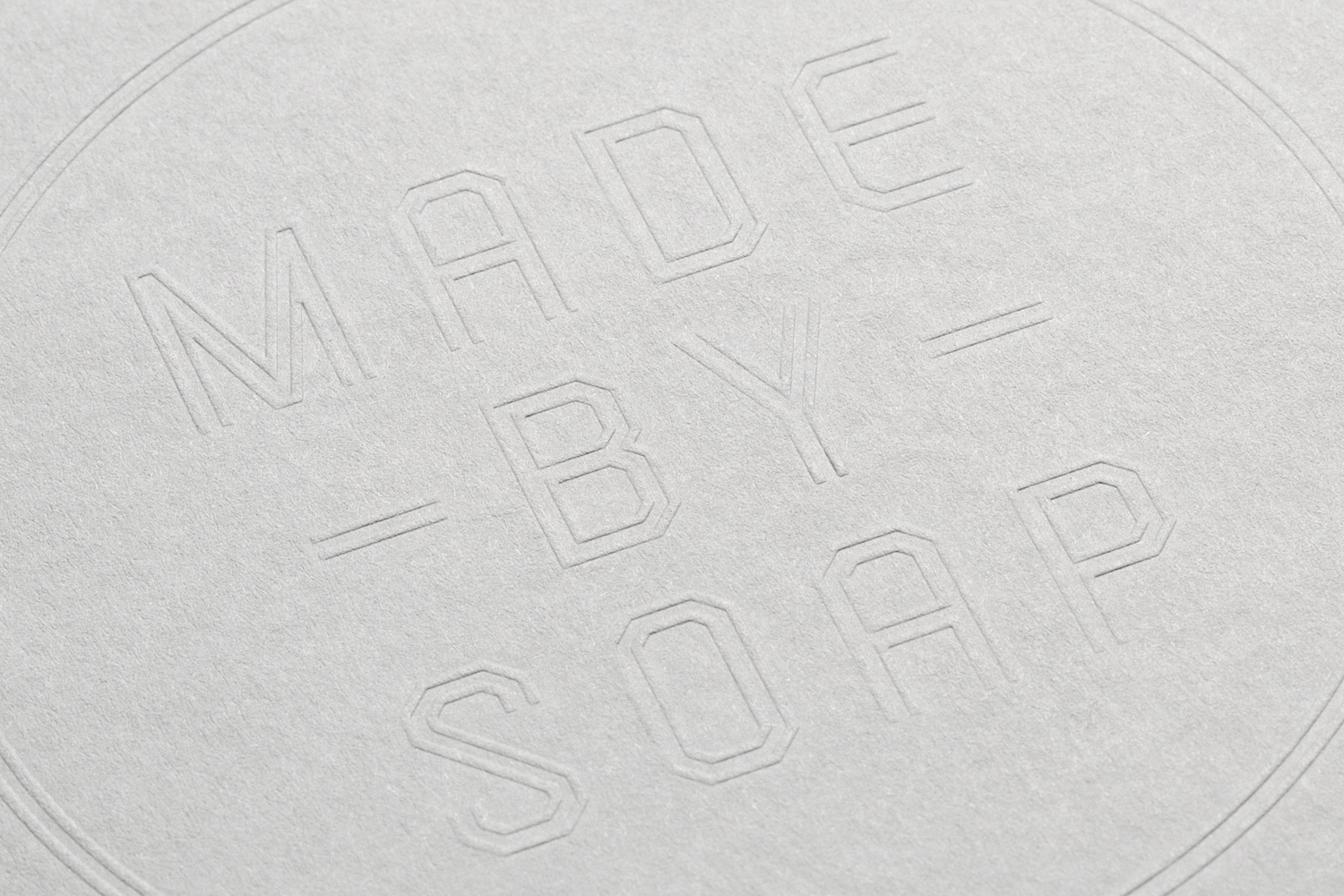 Close-up of debossed typographic logo text reading MADE BY SOAP on a textured light grey paper background.