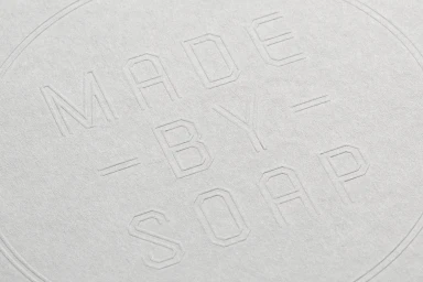 Close-up of debossed typographic logo text reading MADE BY SOAP on a textured light grey paper background.