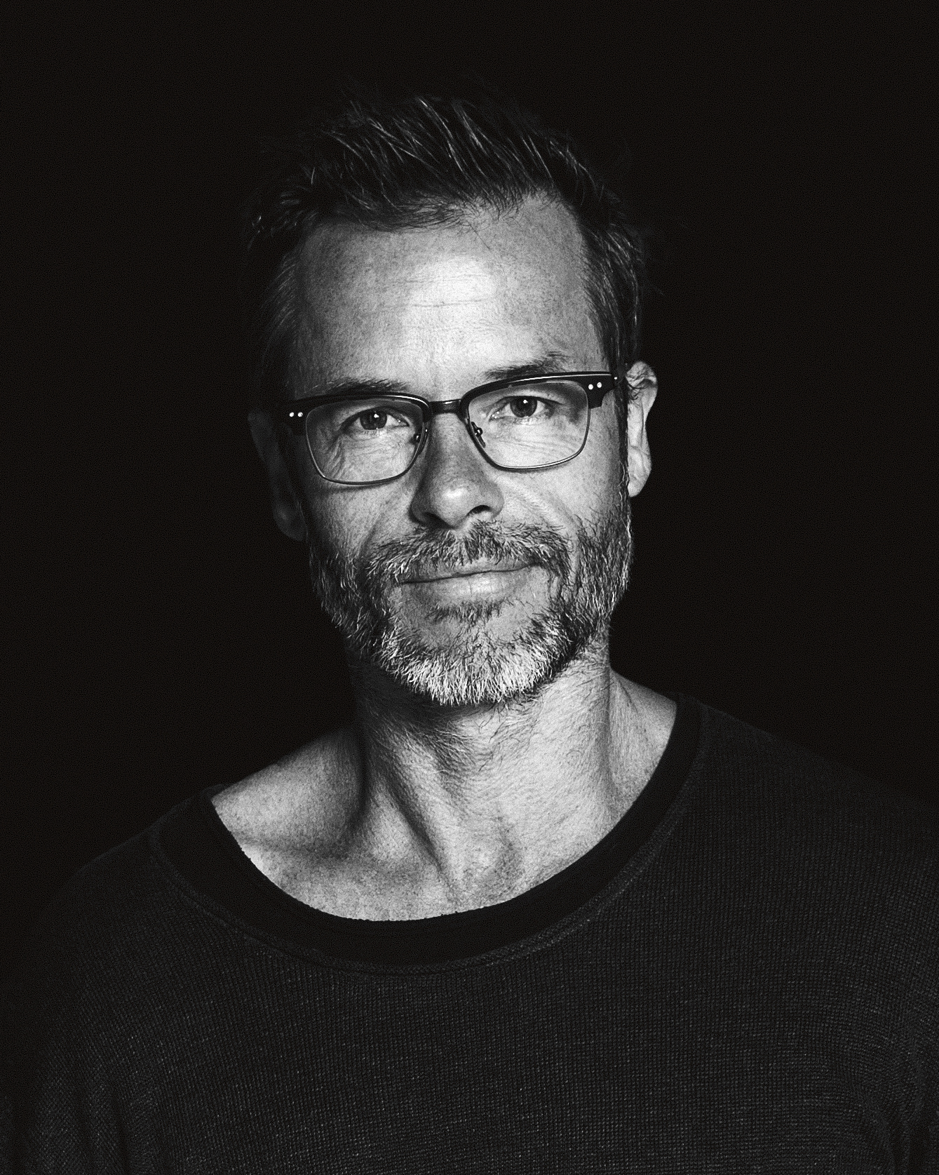Black and white close-up portrait of a man with short hair and glasses wearing a dark sweater against a black background.