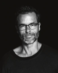 Portrait of actor Guy Pearce wearing glasses and a dark crewneck shirt against a black background by Andrew J Morley