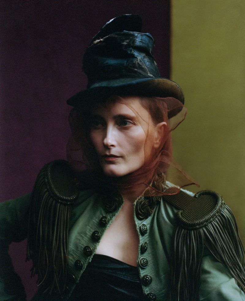 Woman in a dark green coat with tassel epaulettes and a dark hat with netting stands in front of a two-tone background.
