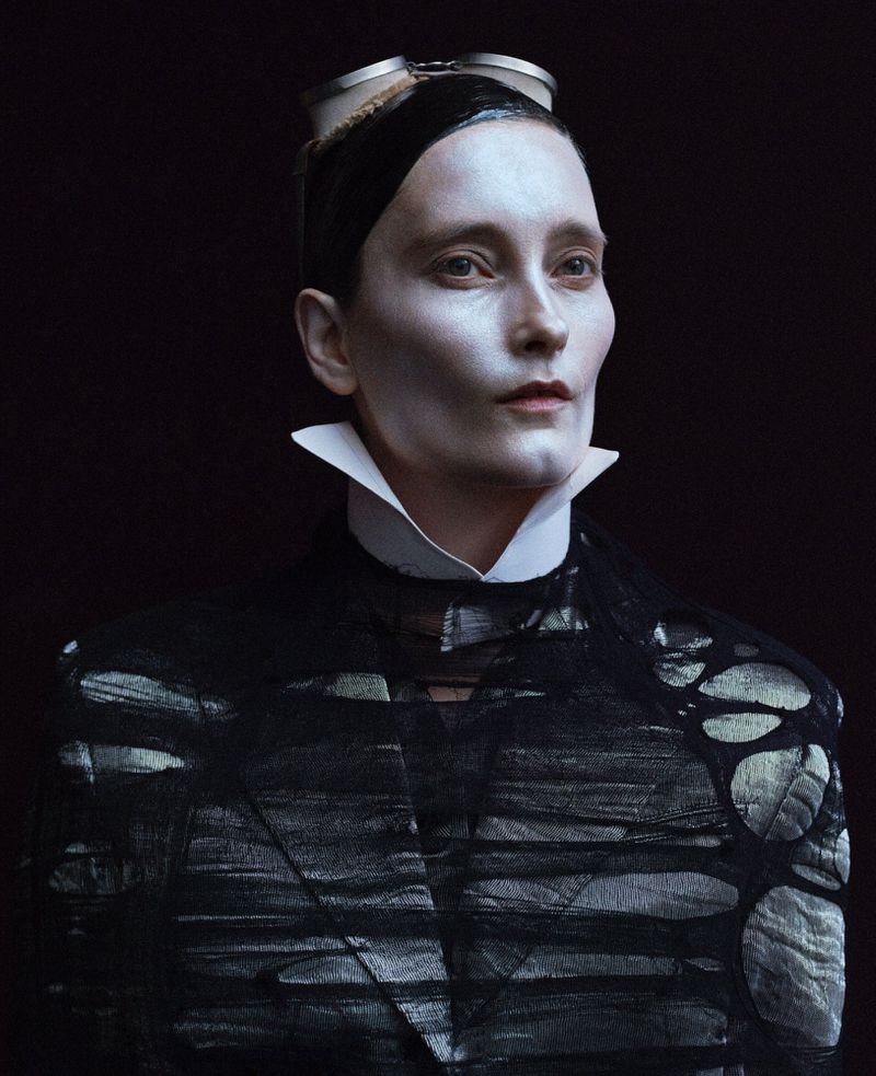 Close-up of a person with pale makeup, wearing a black jacket and goggles, against a dark background. High-fashion, beauty shot.