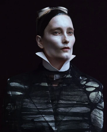 Close-up of a person with pale makeup, wearing a black jacket and goggles, against a dark background. High-fashion, beauty shot.