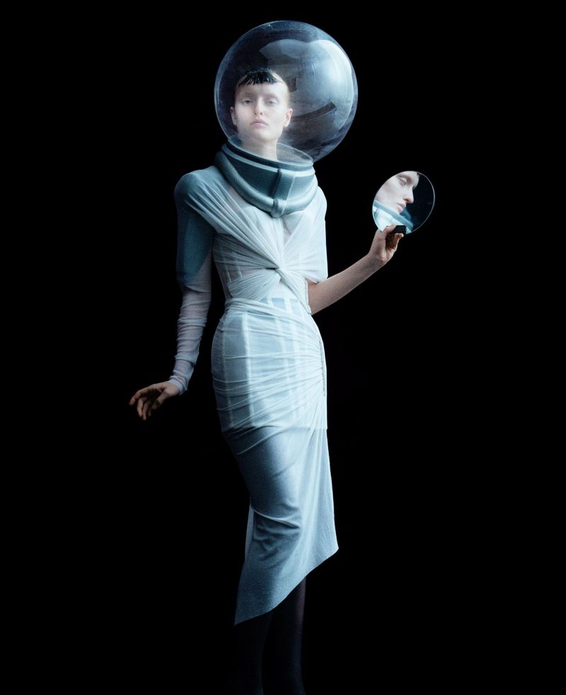 Elegant woman wearing futuristic dress and clear space helmet, holding mirror, isolated on black background in a fashion style.
