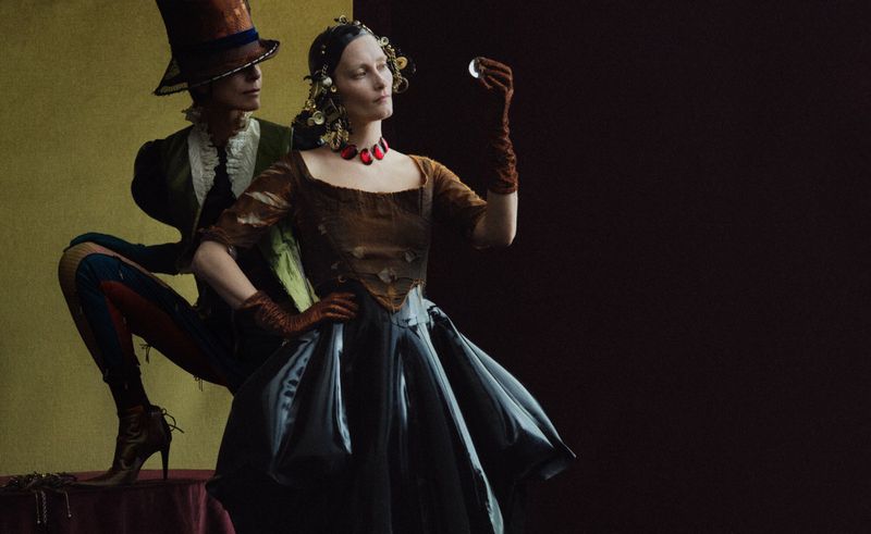 Fashion photography featuring a woman in vintage dress holding an orb and a man in a top hat in a stylized, theatrical composition.