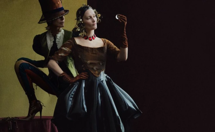 Fashion photography featuring a woman in vintage dress holding an orb and a man in a top hat in a stylized, theatrical composition.