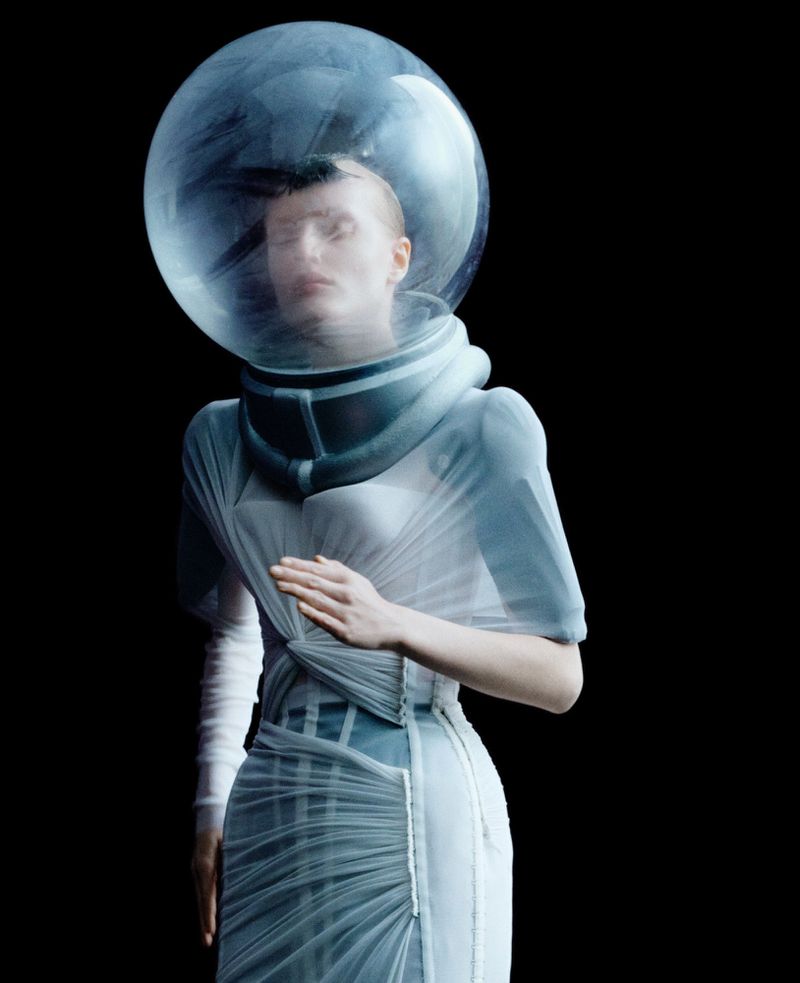 Woman in a light blue dress and astronaut helmet poses against a black backdrop, creating a unique blend of fashion and futuristic design.