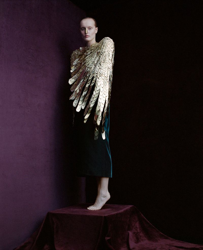 Fashion photo of a woman in a dark green velvet dress and golden feathered wing-like sleeves standing on a draped velvet platform.