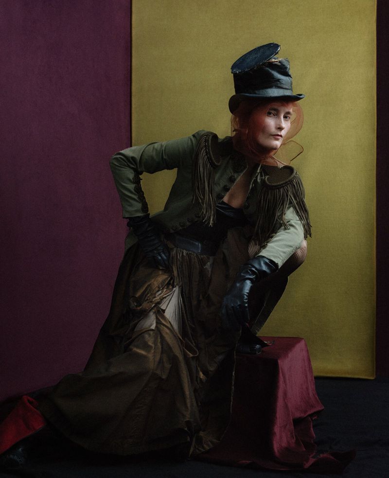 Woman in military-style jacket, top hat, and gown poses in front of a two-tone background with a serious facial expression.