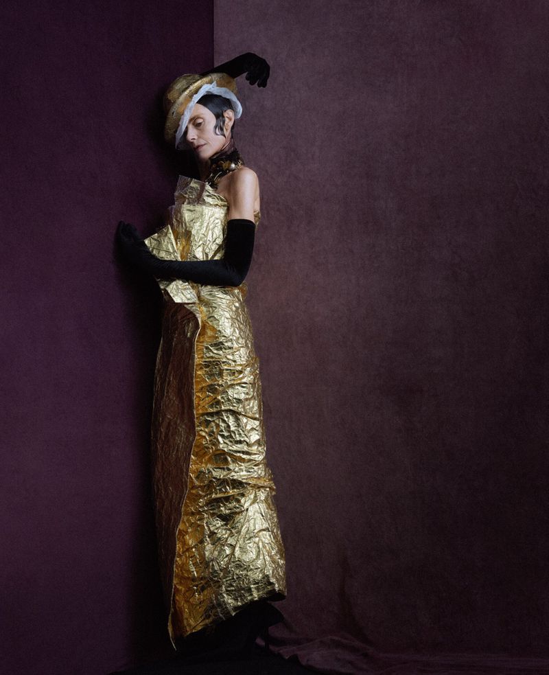 Fashion portrait of a person in a gold dress and straw hat with a feather, posing against a dark burgundy-colored wall, in a studio setting.