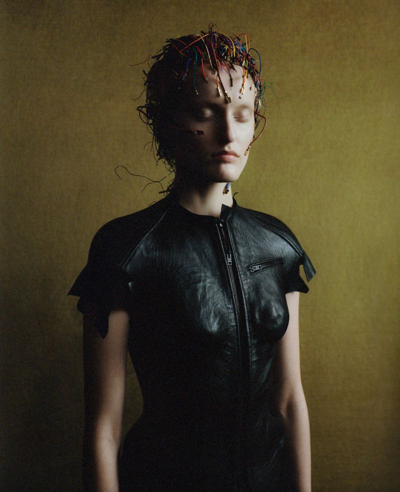 Woman in a leather dress with wires on her head, set against a textured gold background in a studio portrait with a surreal, avant-garde aesthetic.