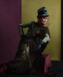 Model in olive green jacket with fringe and black top hat posing against gold and plum panels for Acne Paper