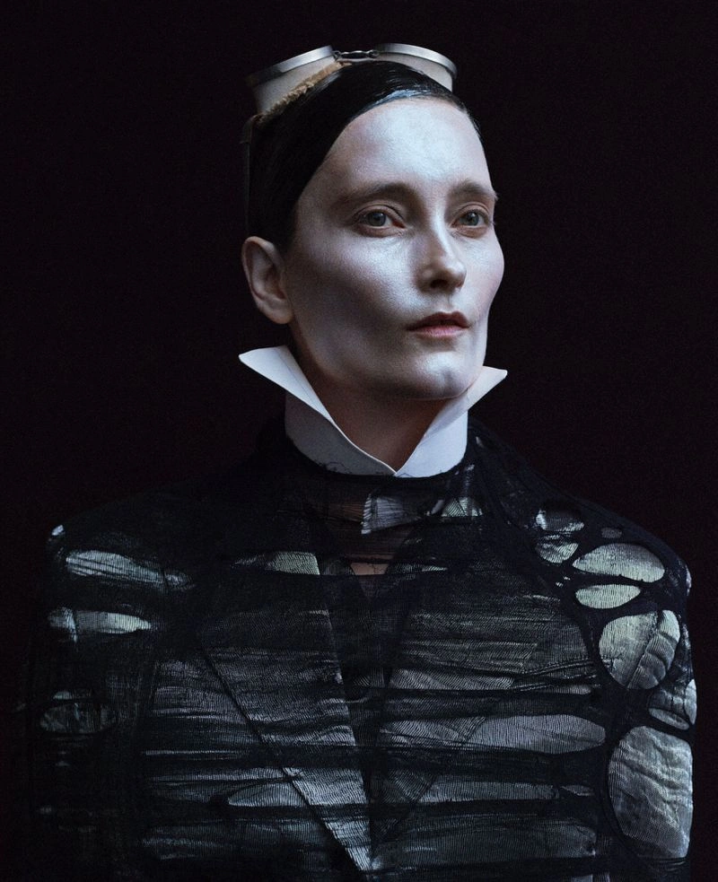 Portrait of a model wearing goggles, white collar, and dark distressed jacket against dark background