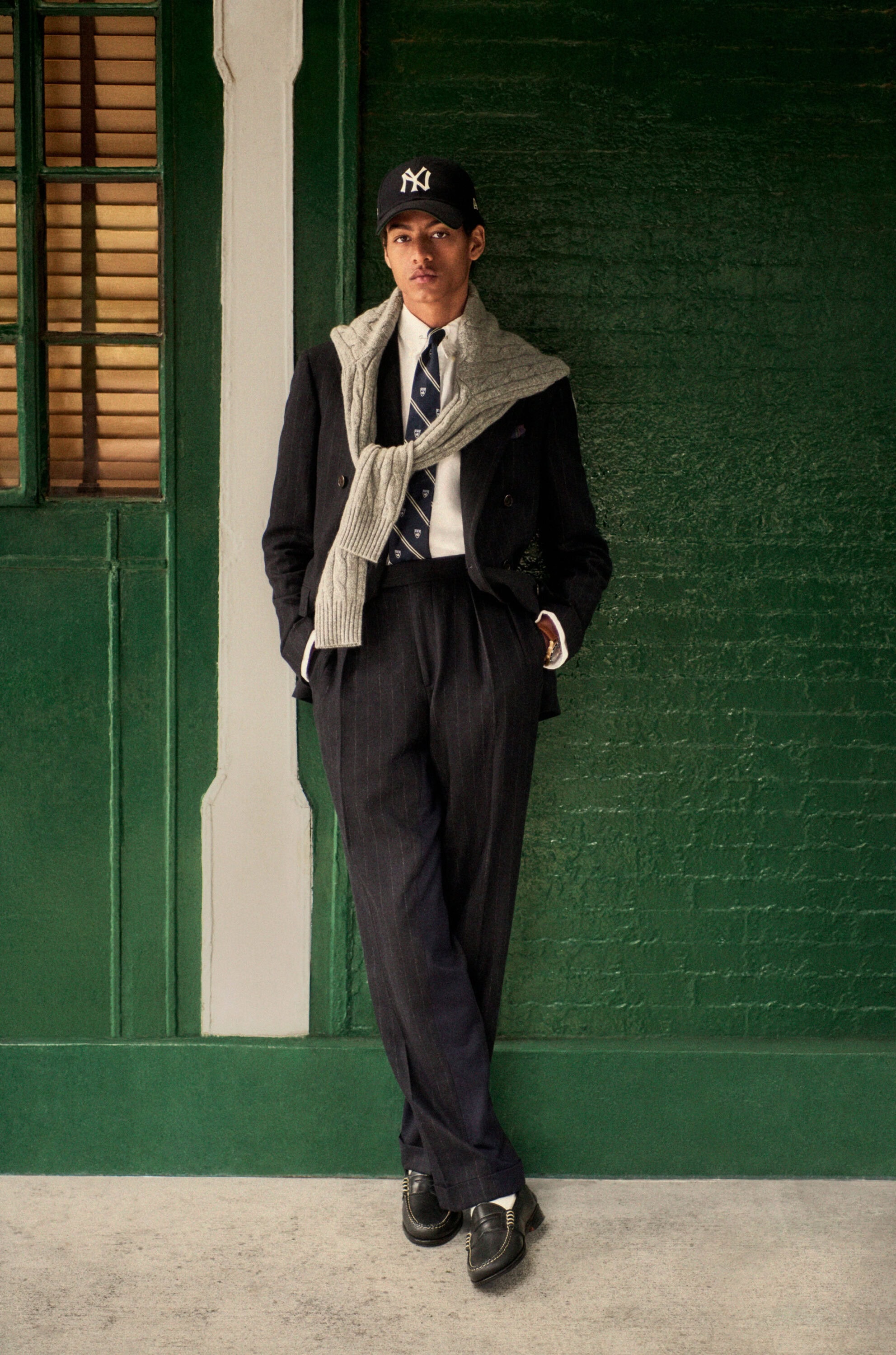 Young man leaning against a green brick wall wearing a dark suit, tie, baseball cap, and a cable-knit sweater tied over his shoulders