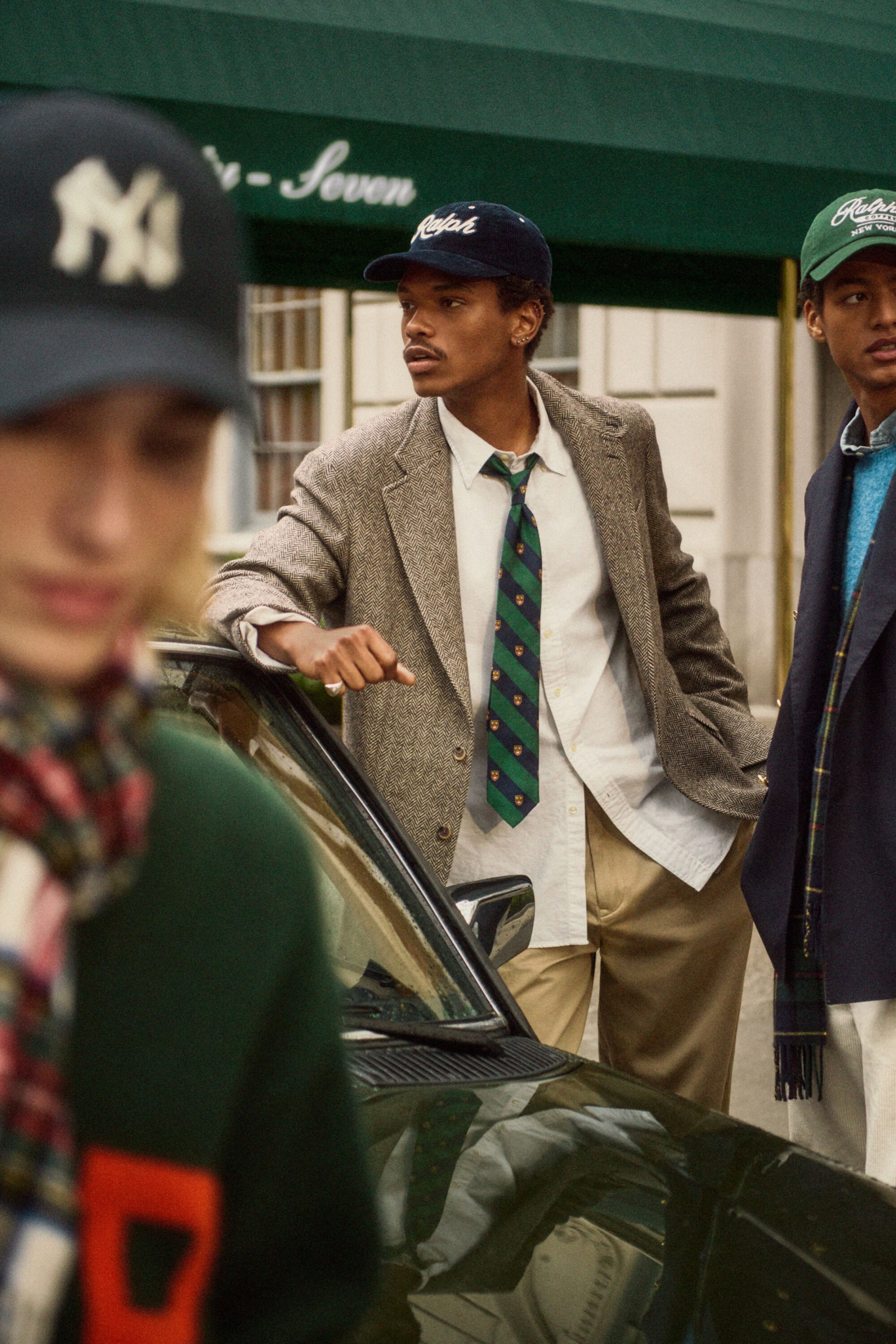 Three young men posing outdoors in layered tailored clothing next to a dark car with a green awning overhead