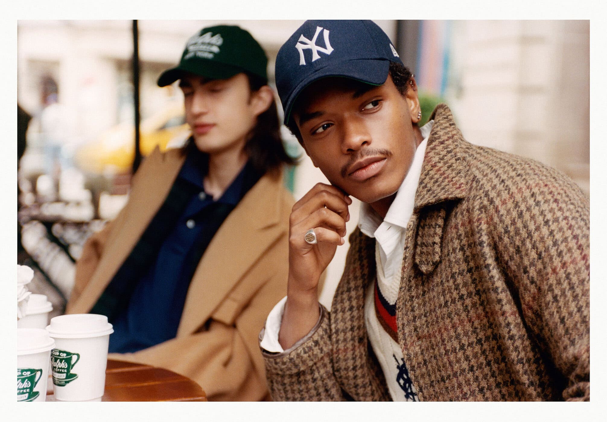 Two male models at an outdoor café table wearing baseball caps and classic tailored outerwear