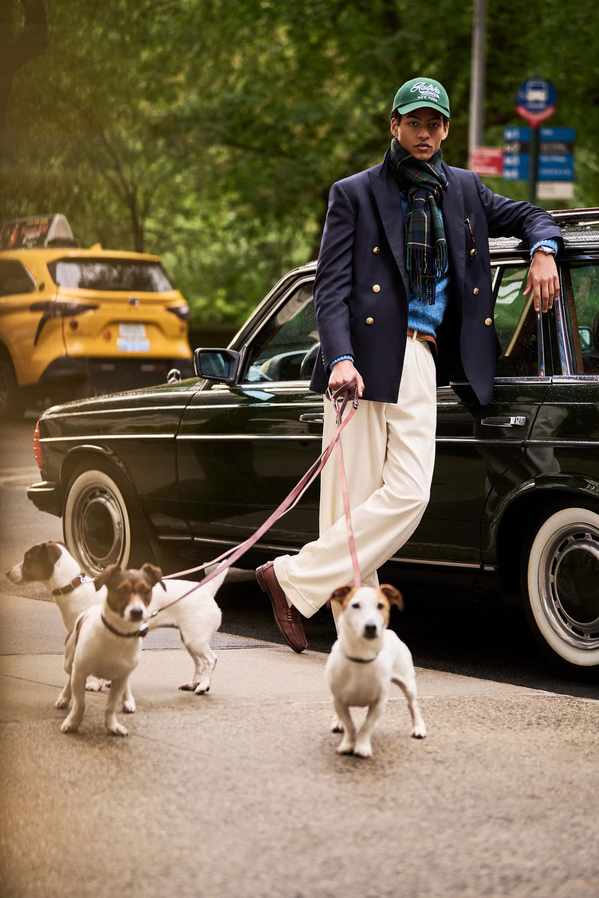Model leaning against a vintage car holding three dogs on leashes on a city street