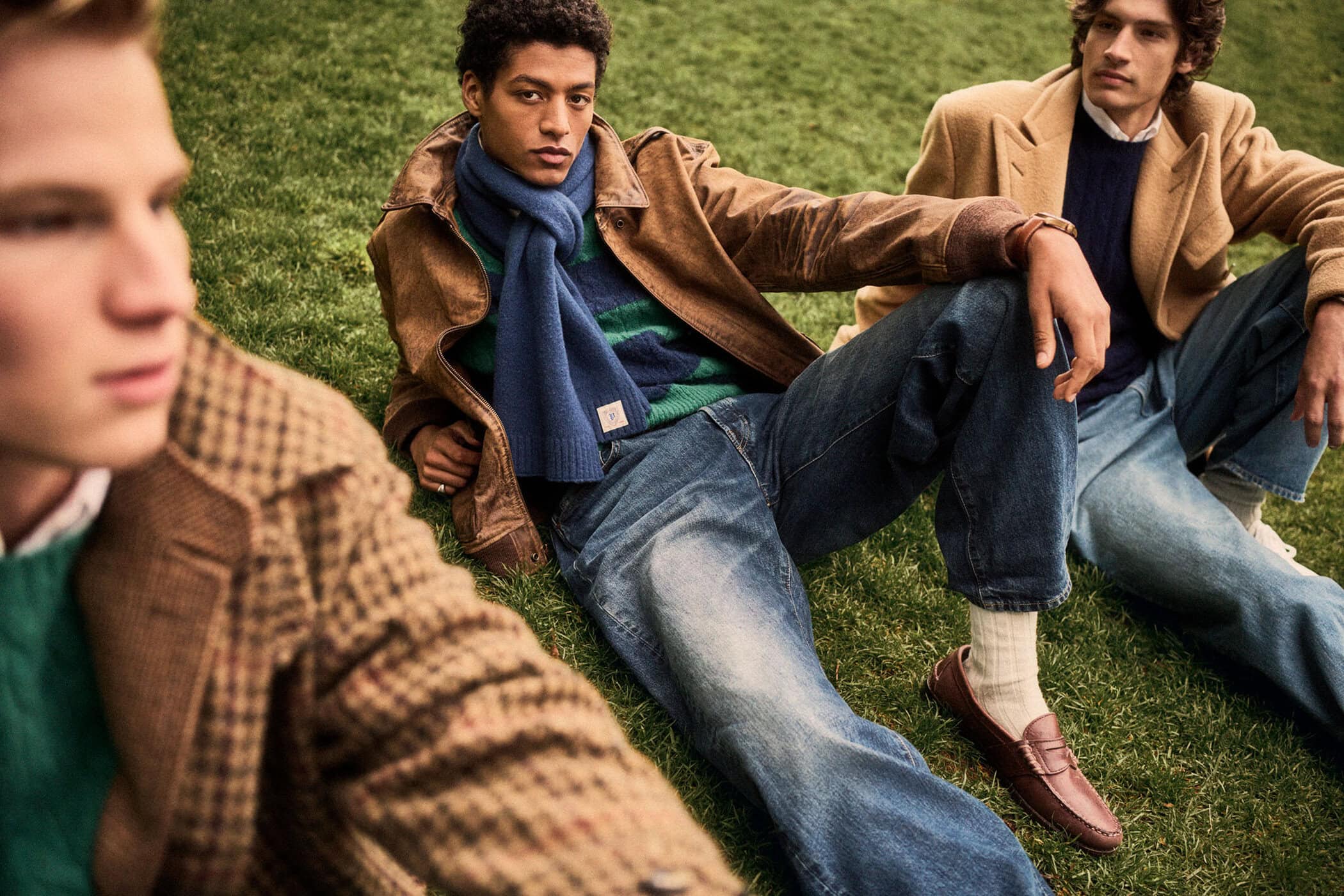 Three young men sitting outdoors on a grassy lawn wearing casual fall outerwear, denim jeans, and knit sweaters.