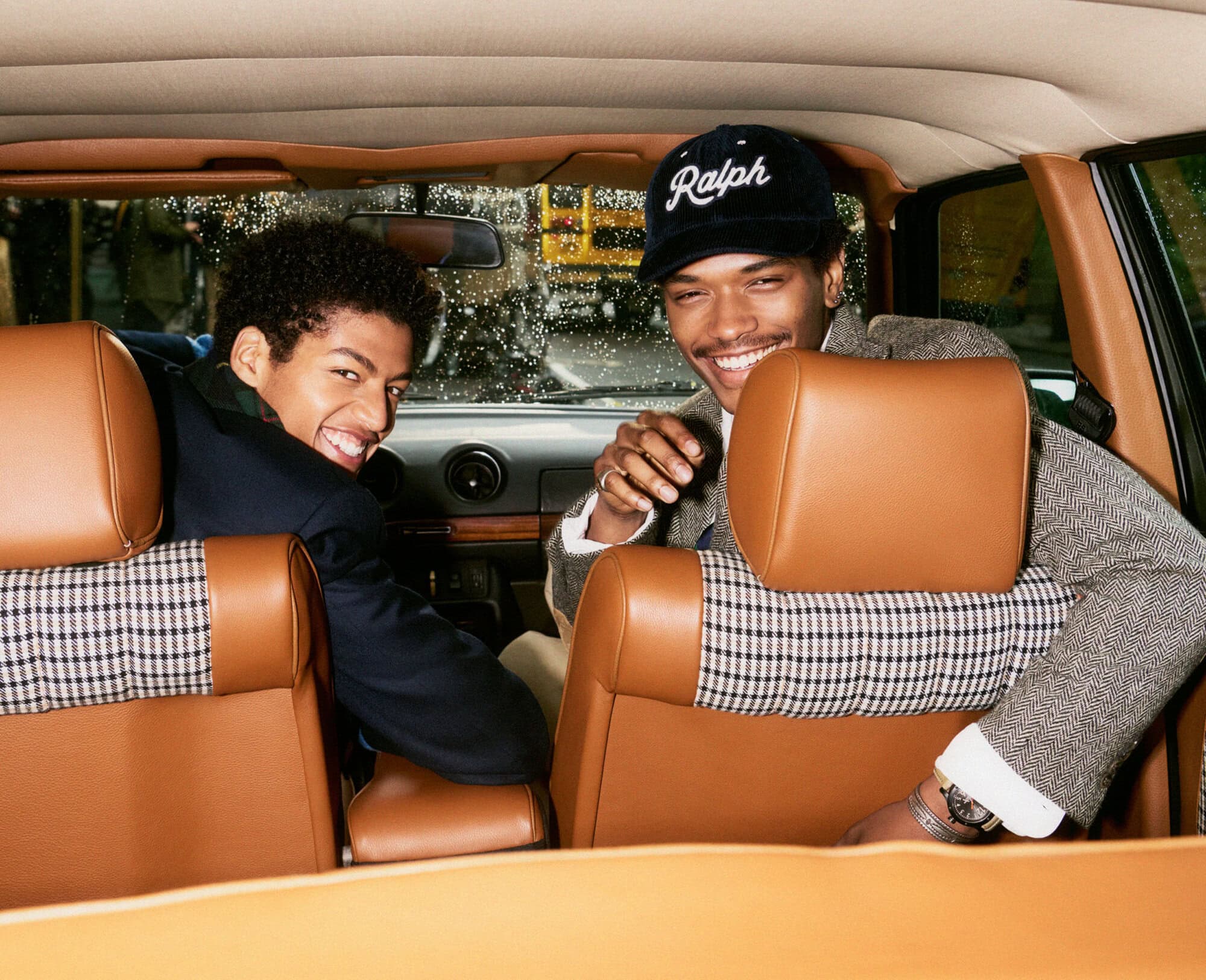 Two smiling male models sitting inside a car with brown leather interior wearing tailored jackets and a baseball cap
