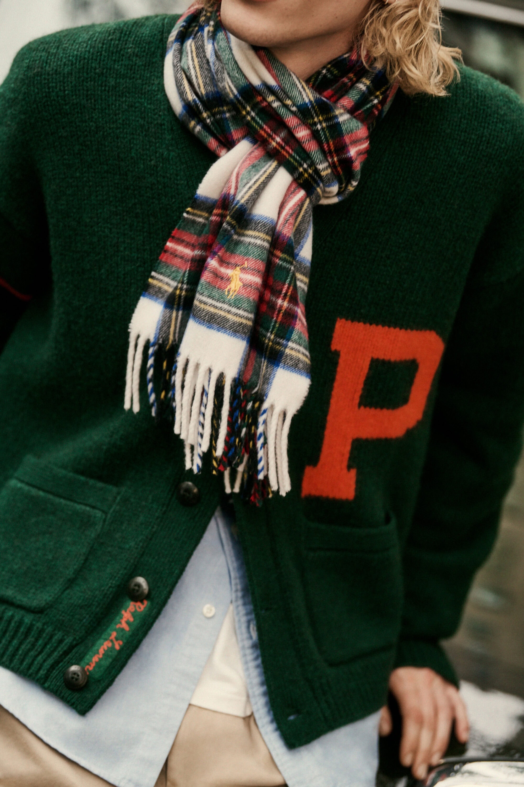 A man wearing a green cardigan with a red letter P and a plaid fringed scarf for Polo Ralph Lauren fall 2025 campaign.