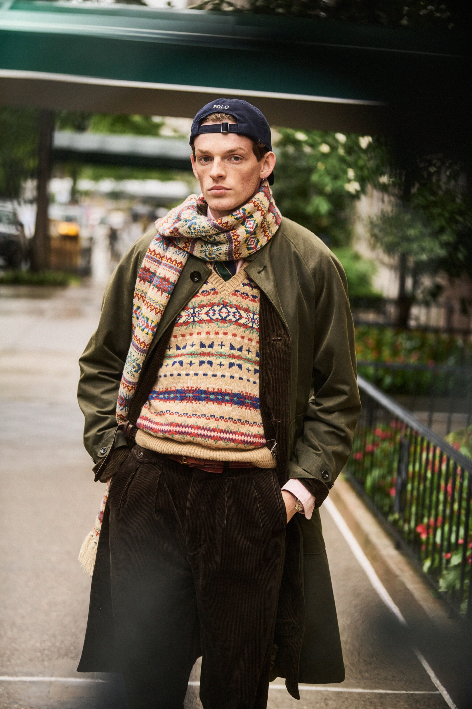 Young man in an olive coat, patterned scarf, and baseball cap standing outdoors on a city sidewalk