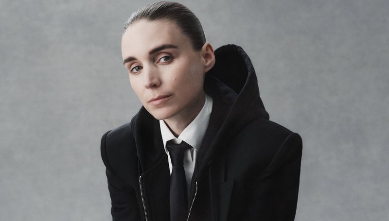 A young adult woman with fair skin and slicked-back brown hair poses indoors wearing a black suit and white dress shirt with a black tie.
