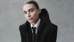 Portrait of Rooney Mara wearing a black hooded jacket, white shirt, and tie against a grey textured studio backdrop.