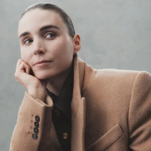 Portrait of Rooney Mara resting chin on hand, wearing a camel blazer and dark shirt against a grey textured studio background.