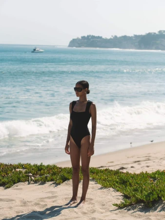 Woman wearing a black swimsuit and sunglasses standing on a sandy beach with ocean waves in the background.