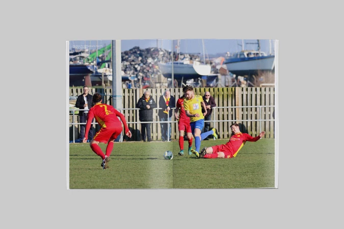 Open magazine spread displaying a football match photograph with players in red and yellow kits on a grass pitch.