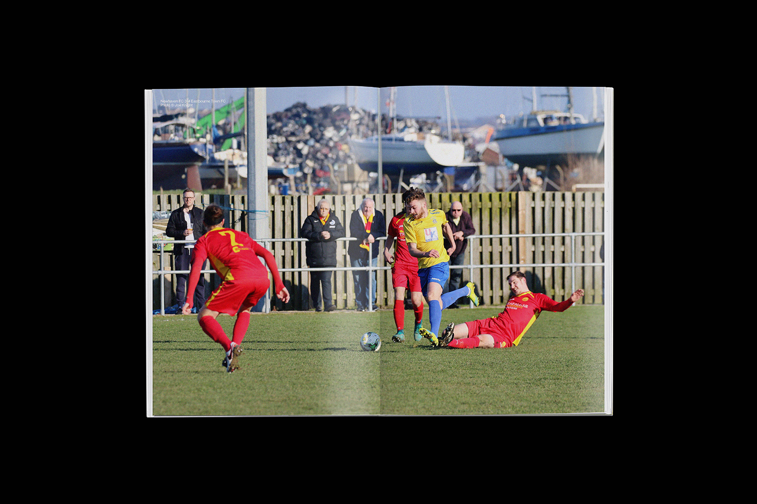 Eastbourne Town FC football players in action during a match on a grass pitch with a marina background