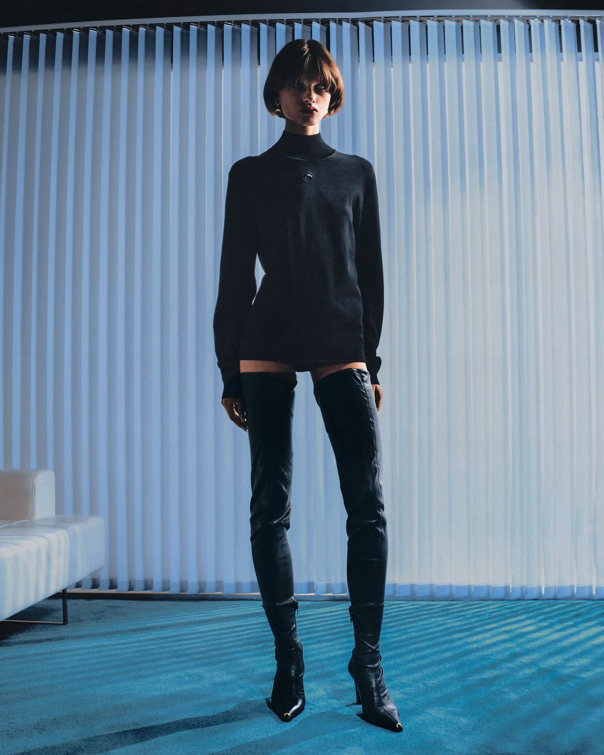 Model with short hair wearing a black turtleneck and thigh-high boots in a room with blue vertical blinds.