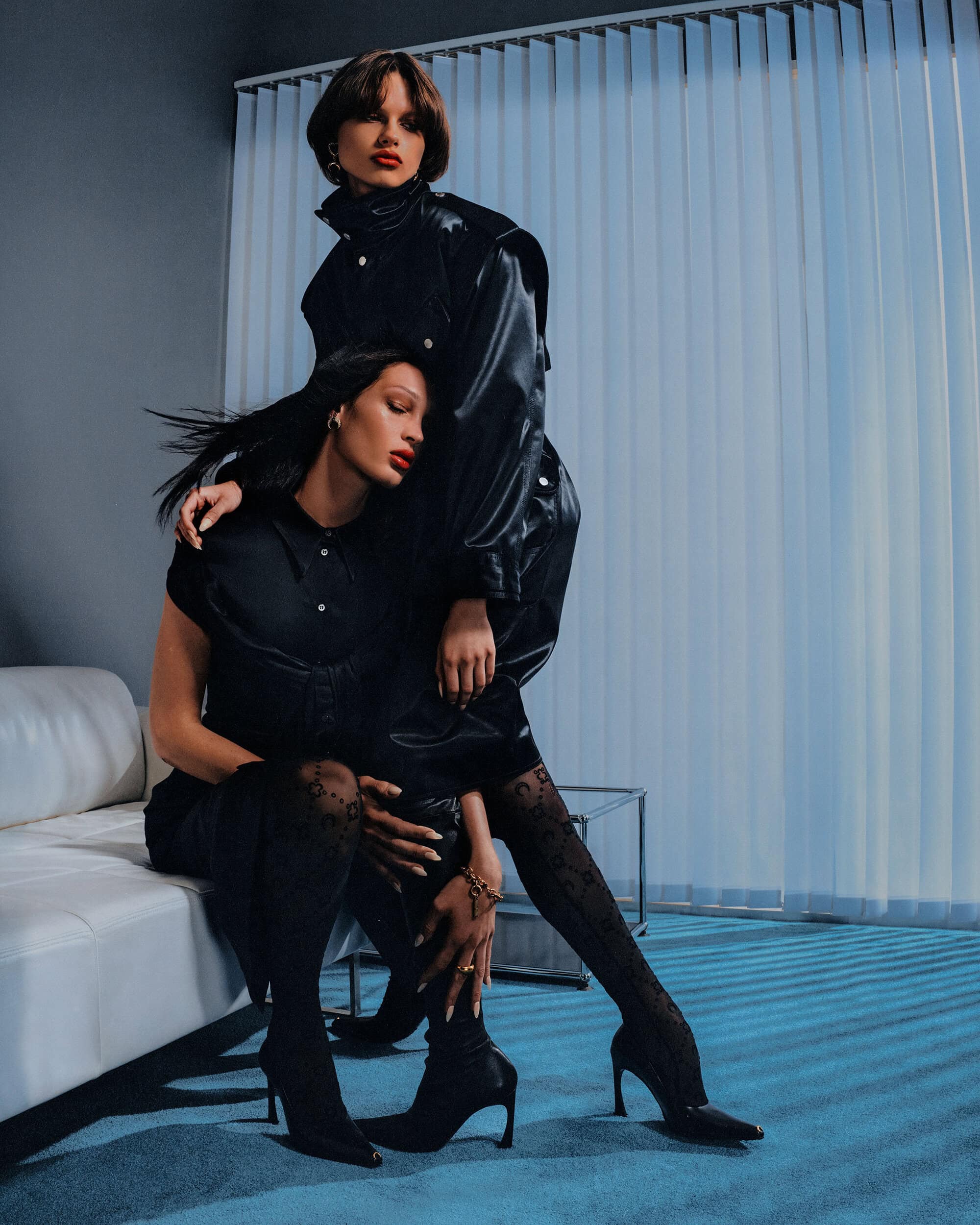 Two female models in black outfits posing together with a white sofa against a blue vertical-blind background.