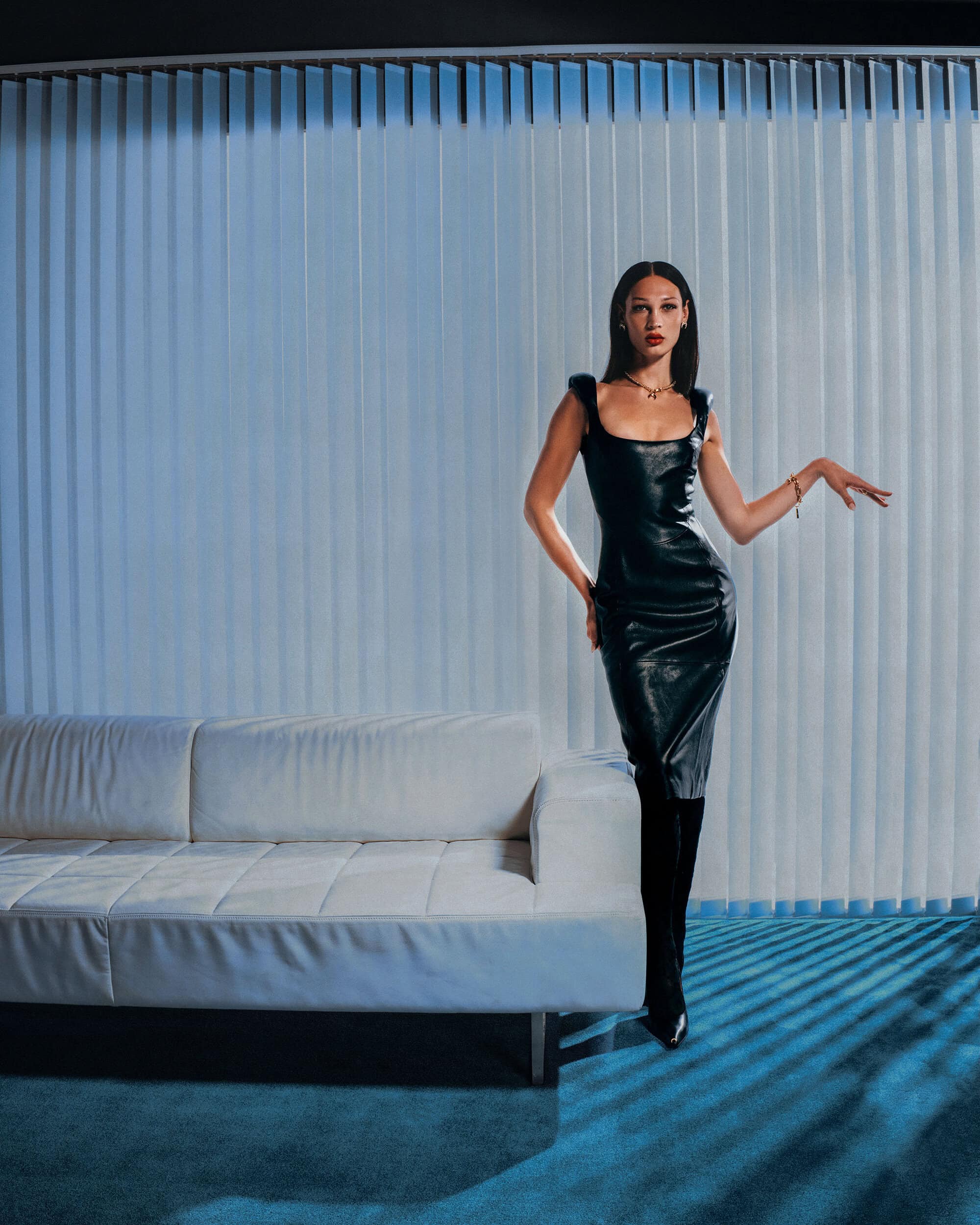 Model in a black leather dress standing next to a white sofa against vertical blinds