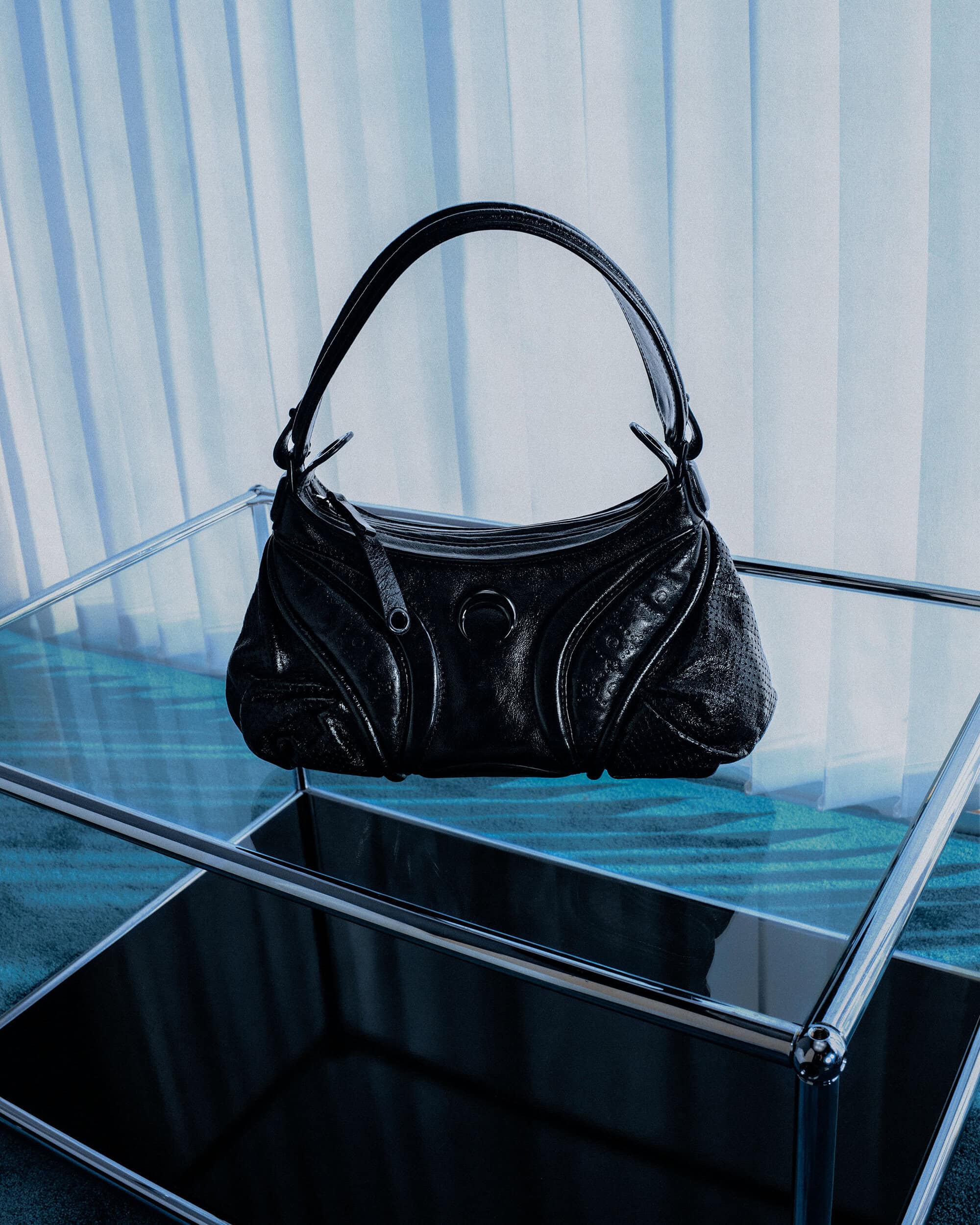Black leather handbag resting on a modern glass and chrome table against draped white curtains.