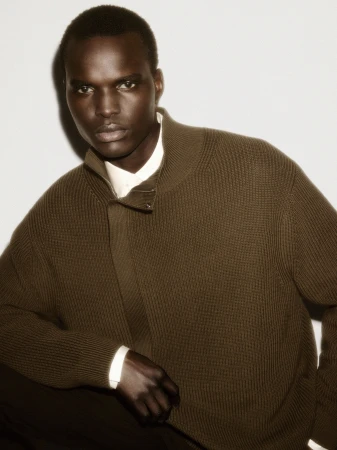Man wearing an olive green zip-up knit sweater over a white collared shirt against a plain light studio background.