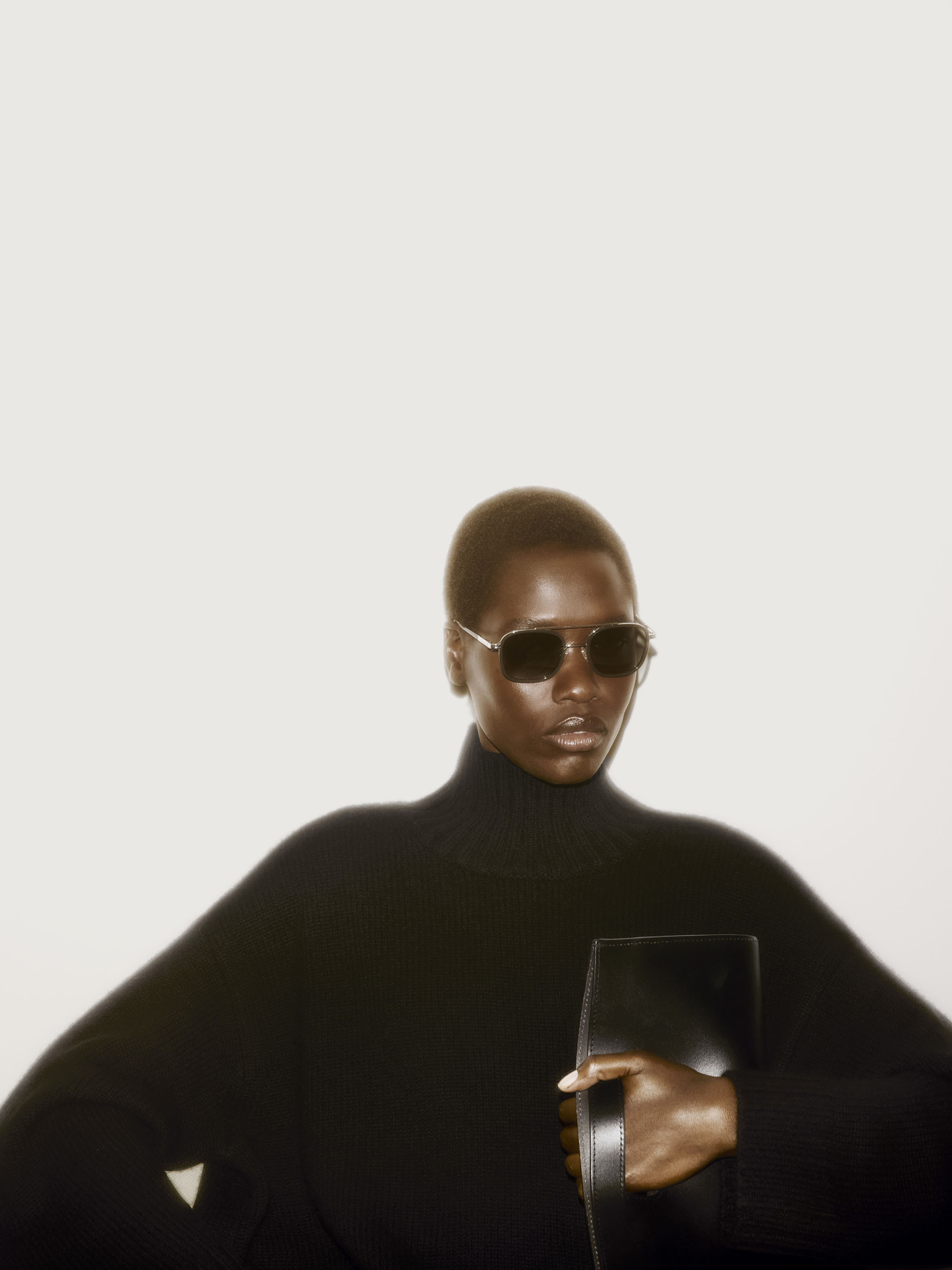 Model in dark sunglasses and black turtleneck holding a leather clutch against a plain white background for COS