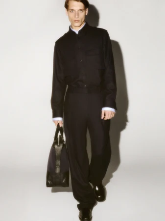 Man wearing a dark button-up shirt and trousers holding a black handbag against a seamless white background