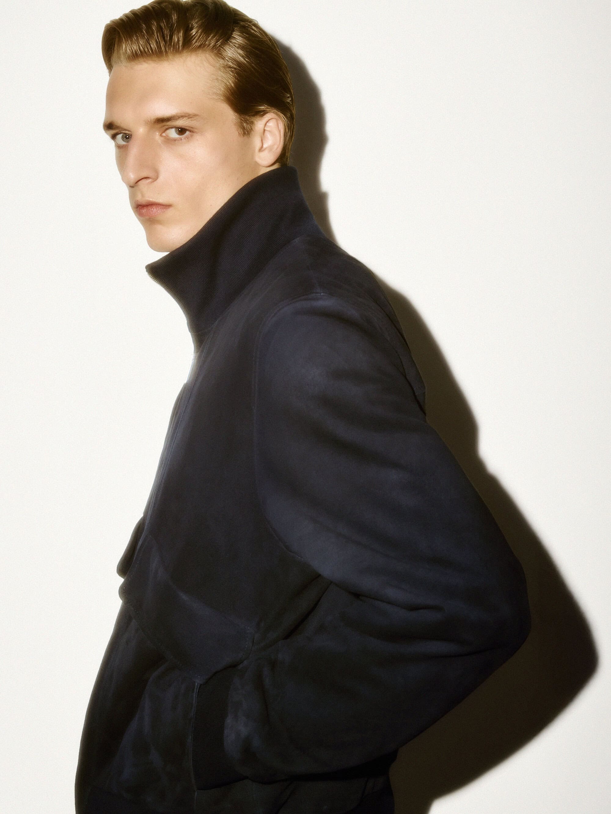 A man wearing a dark high-collar jacket against a stark white wall with a sharp shadow cast behind him.