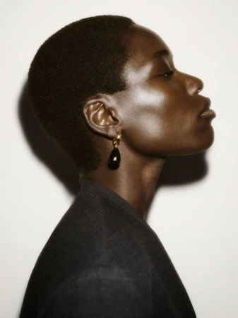 Profile portrait of a model with short dark hair wearing a black top and a single teardrop earring against a plain white background.