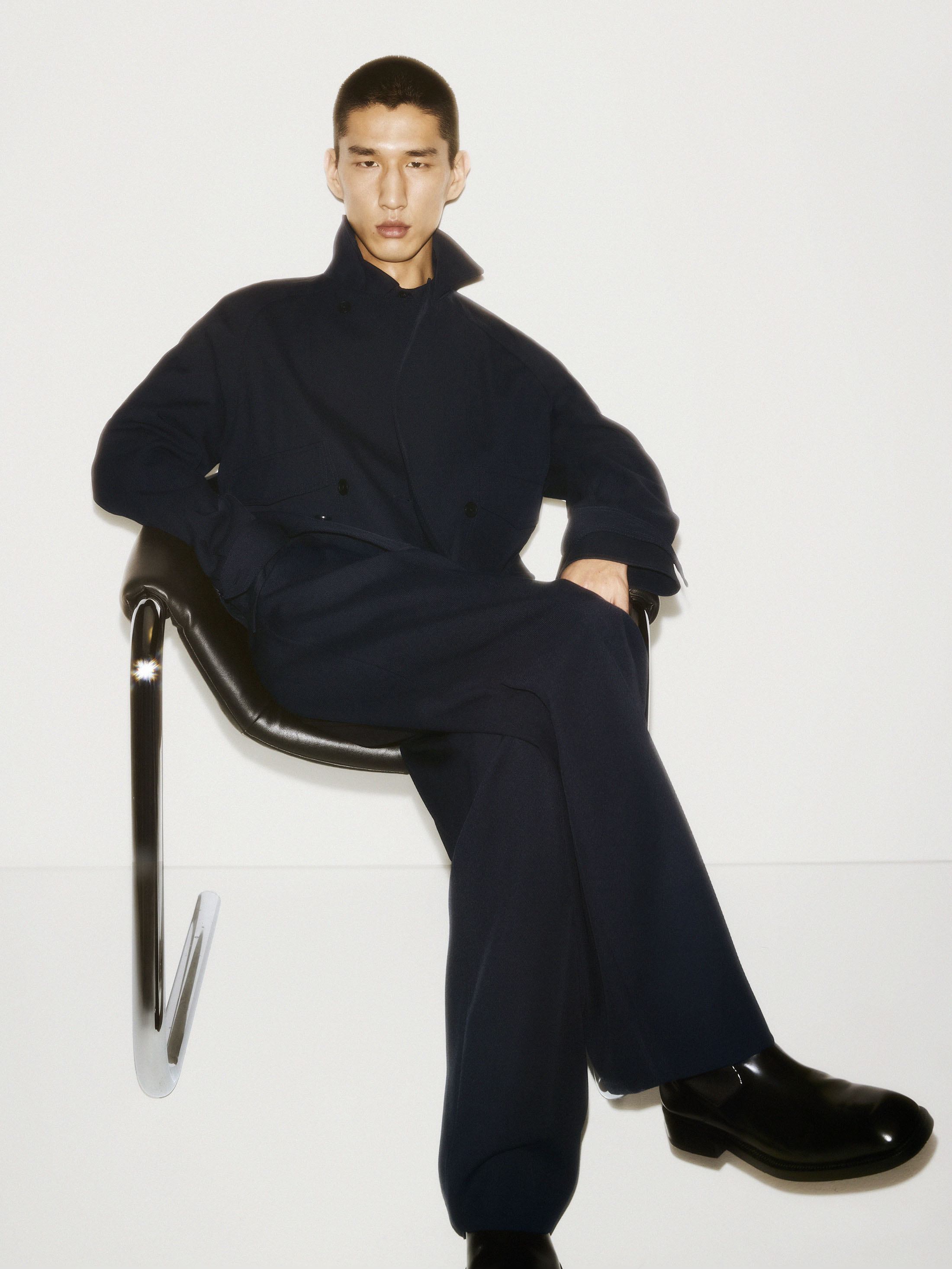 Man seated on a modern black chair wearing a dark wool coat against a seamless white background for COS